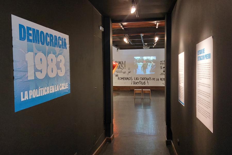 Imagen de un pasillo pintado de negro, donde en la izquierda esta colgado un cartel azul con letras blancas que dice "Democracia. 1983. La política en la calle."