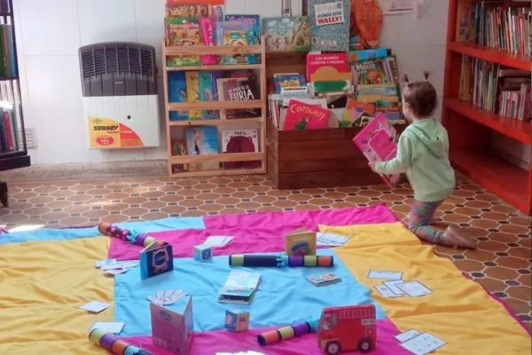 Un espacio con una manta y juegos de tela sobre el piso, hay un niño pequeño con un libro, también bibliotecas bajas a la altura de las infancias