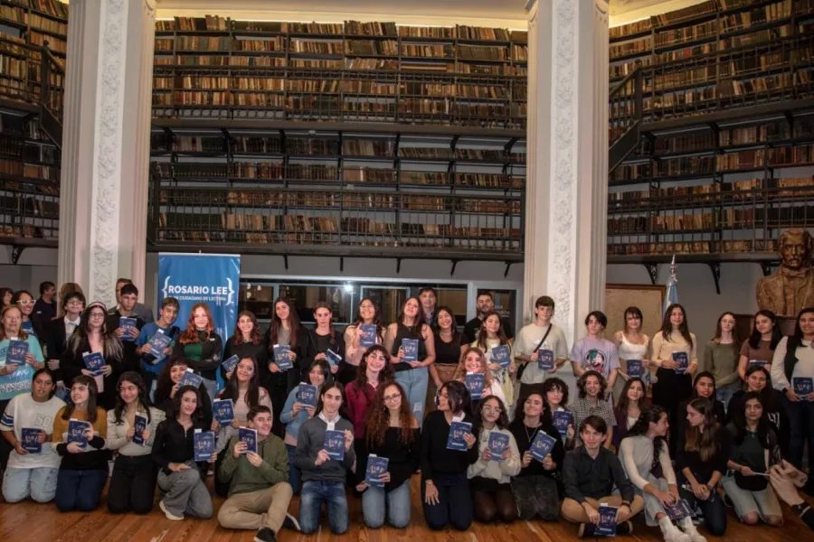 Foto de muchos chicas y chicos con libros en la mano, de fondo una gran biblioteca