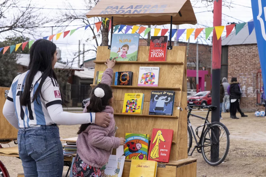 Una chica joven junto a una niña, ambas de espaldas, frente a un carro biblioteca con libros infantiles con colores e ilustraciones