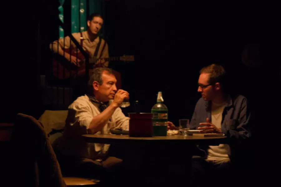 Es la imagen de una escena de una obra de teatro. Hay tres actores: dos sentados tomando bebidas y hablando y de fondo, hay una persona sentada en una escalera tocando la guitarra
