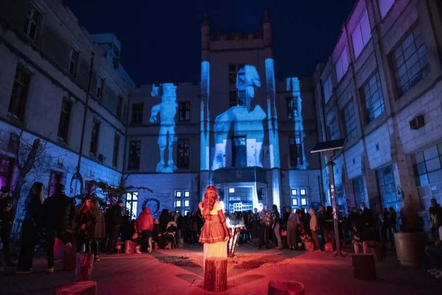 Es la imagen del patio de la facultad de Humanidades, donde en el edificio se proyectan imagenes y hay personas escuchando a la presentadora