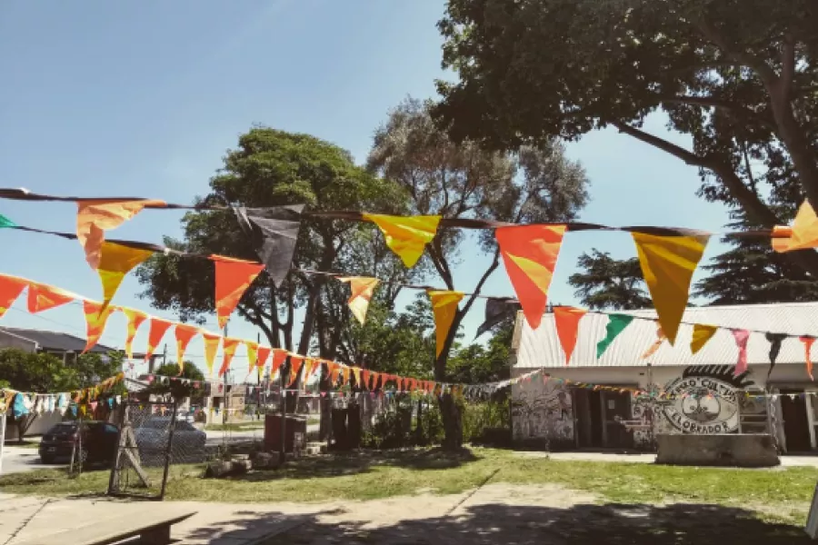 Es la imagen del Centro Cultural El Obrador. Es una fotografía previa a un evento donde se observan banderines de diferentes colores en un espacio verde grande. En el fondo, se observa una casa