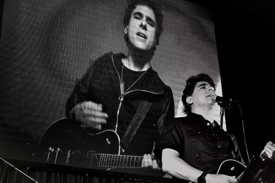 Es la imagen de un músico que está tocando una guitarra eléctrica. De fondo, hay una pantalla gigante donde se refleja lo que hace el músico