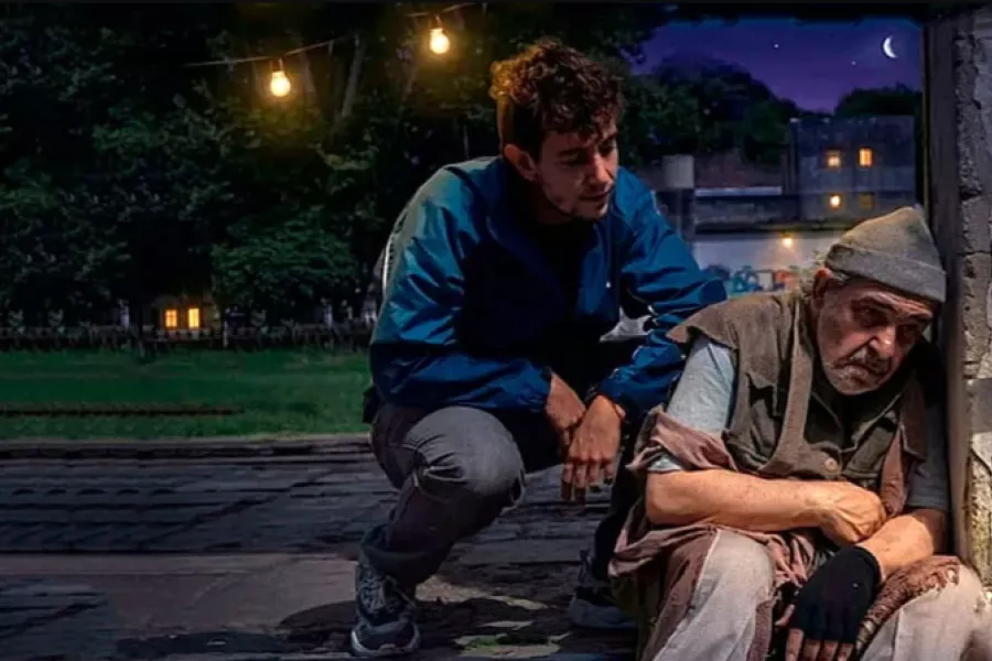 Es una imagen de dos actores caracterizados como sus personajes y realizando una escena: una persona mayor sentada y apoyando la cabeza en una pared y otra persona más joven, de cuclillas, hablándole