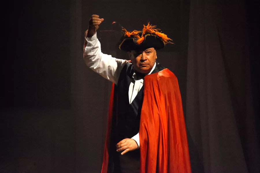 Es la imagen de un actor caracterizado con una vestimenta antigua: un sombrero negro con plumas naranjas, una capa roja, una camisa blanca y un chaleco negro