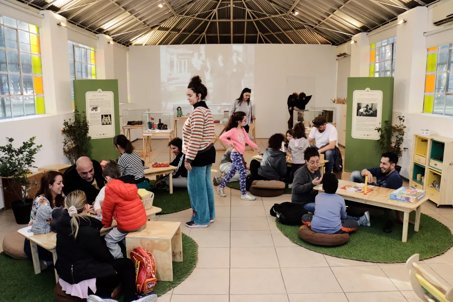 Foto del interior del Espacio Invernadero, en mesas dispuestas para jugar, infancias junta a sus familias disfrutan de distintos juegos de mesa, de pie las coordinadores de la propuesta.