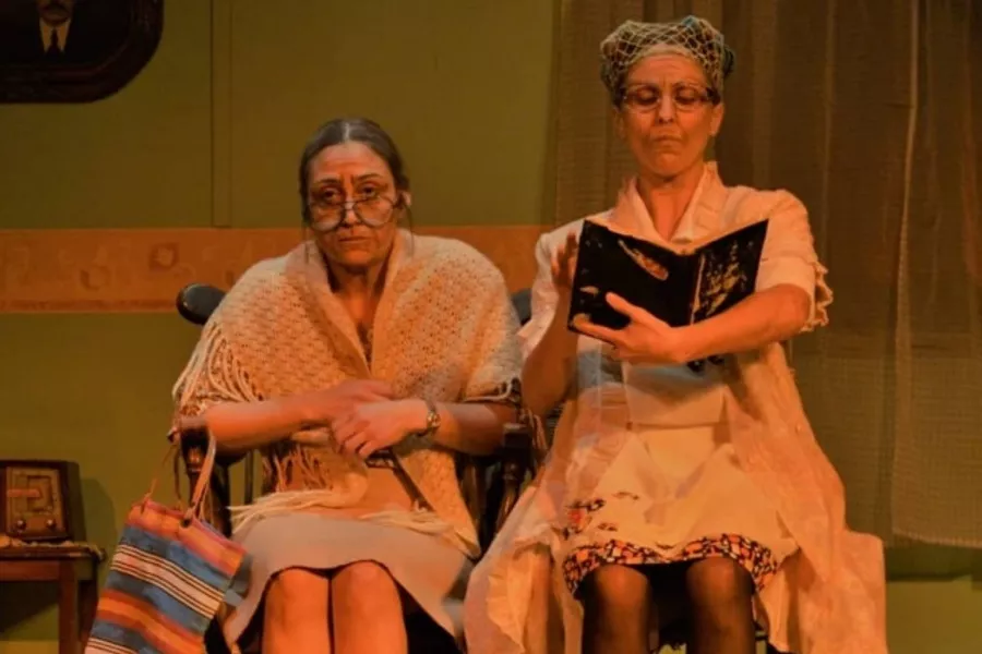 Es la imagen de dos actrices caracterizadas como dos señoras mayores. Una está leyendo un libro y la otra se está agarrando las manos 