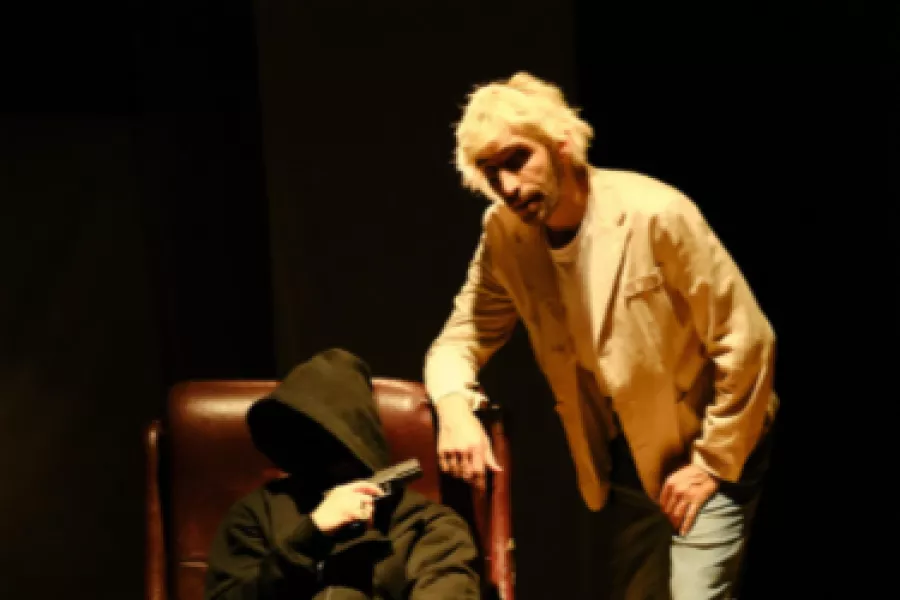 Es la imagen de dos actores caracterizados como sus personajes. Uno está sentado con un arma y el otro está agachado apoyándose en el sillón