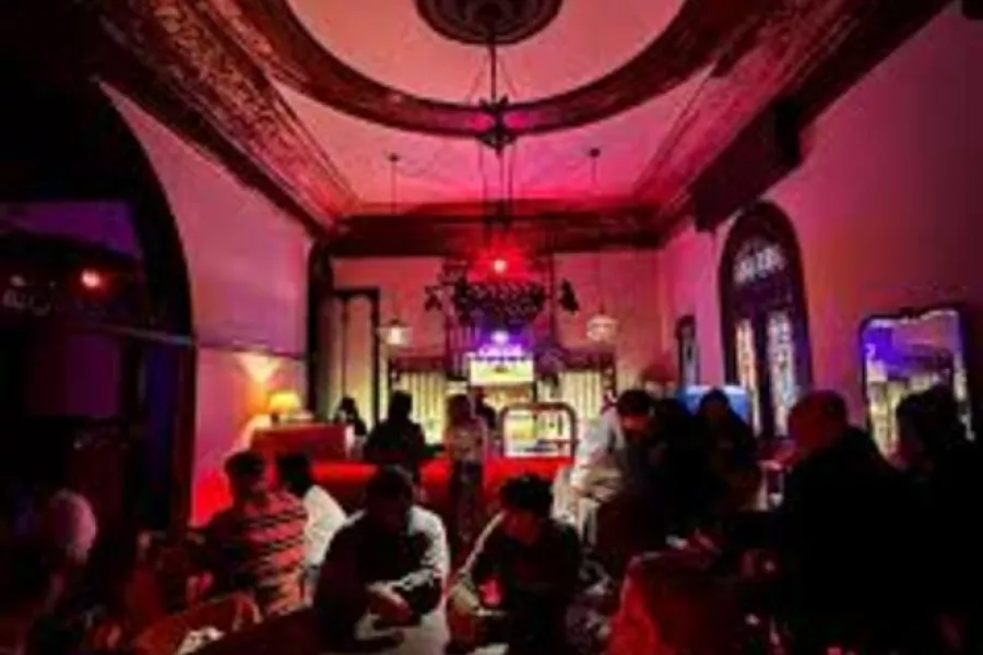 Es la imagen del interior de una casa antigua: presenta un techo alto y una lámpara en el medio. Hay mesas y sillas y personas charlando 