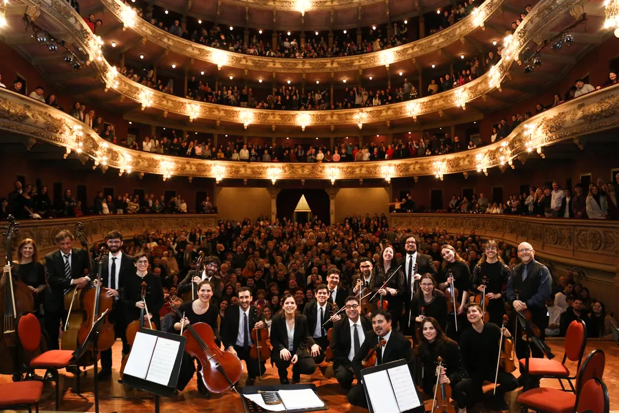 Es la imagen de una orquesta posando en un teatro repleto de personas