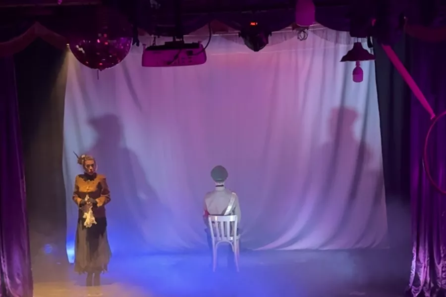 Es la imagen del escenario de Club Fosse, está decorado con cortinas y luces. En el medio hay una actriz caracterizada como su personaje y otro actor sentado de espalda al público
