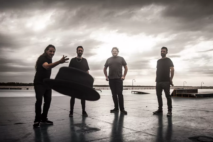 Es la imagen de un grupo de músicos posando para una fotografía, están parados en una playa y uno de ellos está lanzando un sombrero hacia la cámara