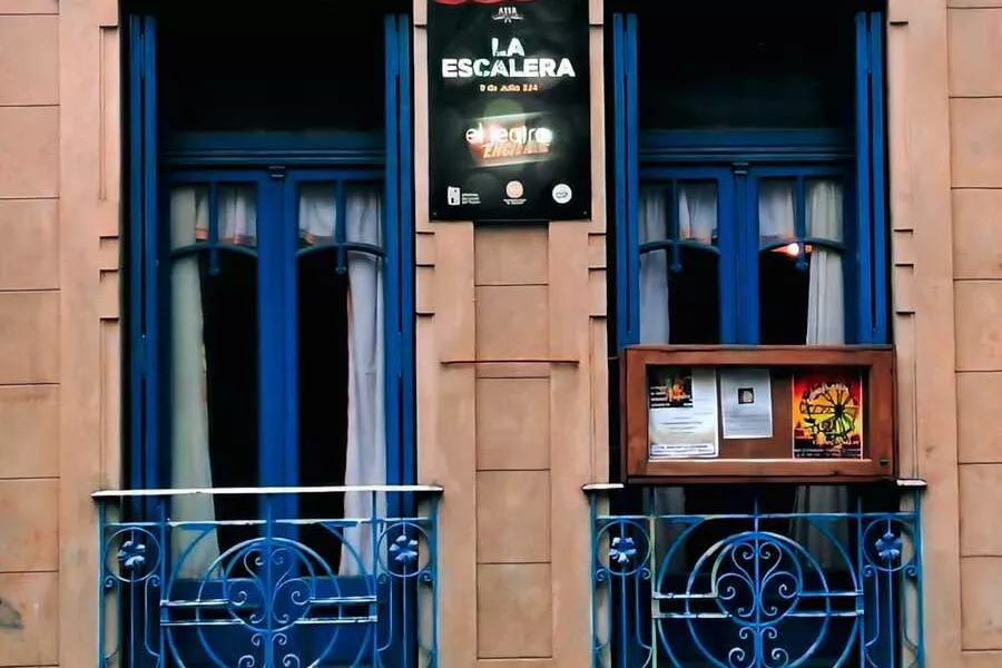 Es la imagen de la fachada del teatro la escalera, es un edificio antiguo con dos ventanas decoradas con cortinas azules. En el medio de ellas, está el cartel del teatro