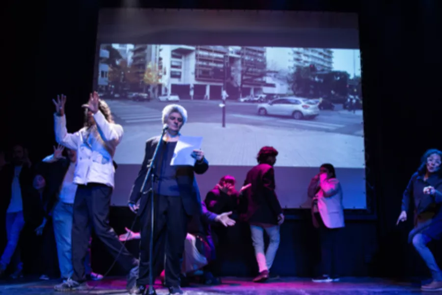 Es la imagen de un escenario donde una persona está leyendo mientras atrás hay artistas haciendo una coreografía