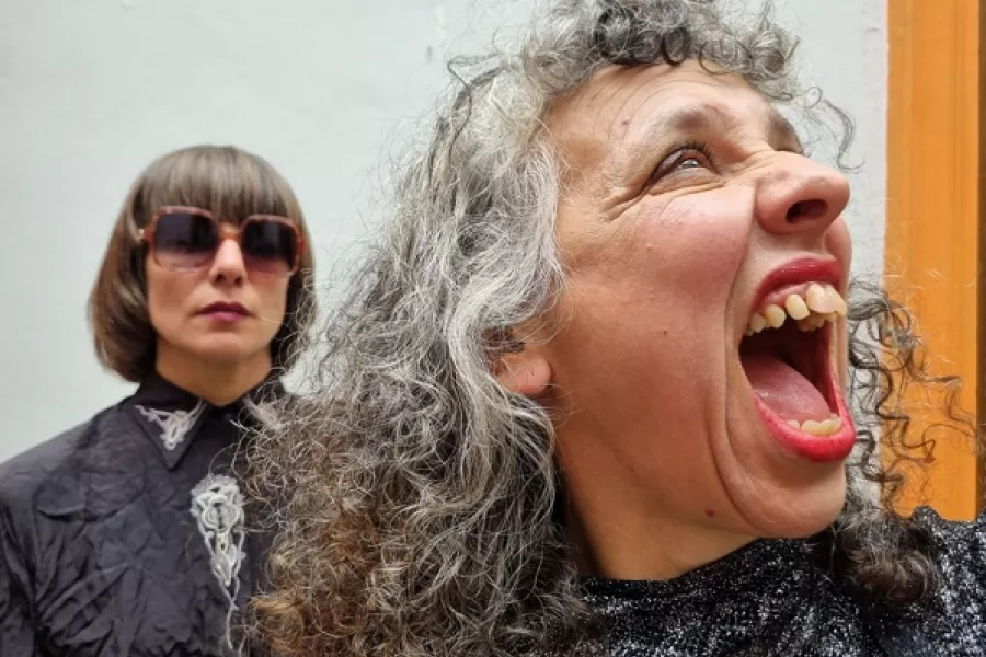 Es la imagen de dos actrices caracterizadas como dos señoras: una está gritando y la otra la está mirando con cara seria y lentes de sol