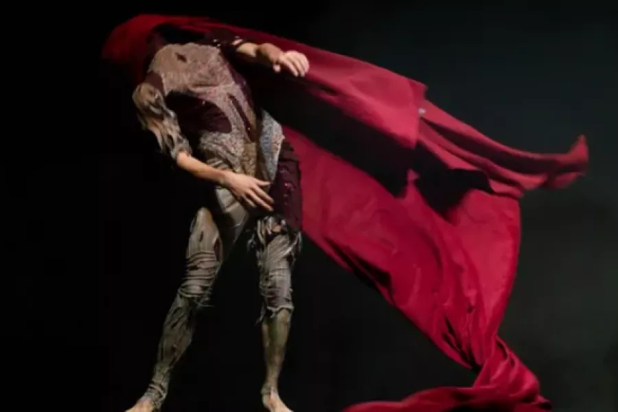Es la imagen de un actor realizando una escena. Tiene un traje de color gris y una capa roja en la cabeza
