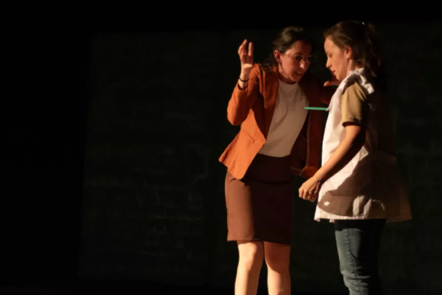 Es la imagen de dos actrices caracterizadas como sus personajes. Una está vestida con un saco naranja y la otra actriz, con un guardapolvo blanco