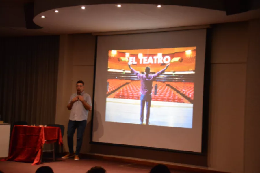 Es la imagen de una presentación de un actor. Está hablando por un microfóno con una pantalla atrás con el título de la presentación