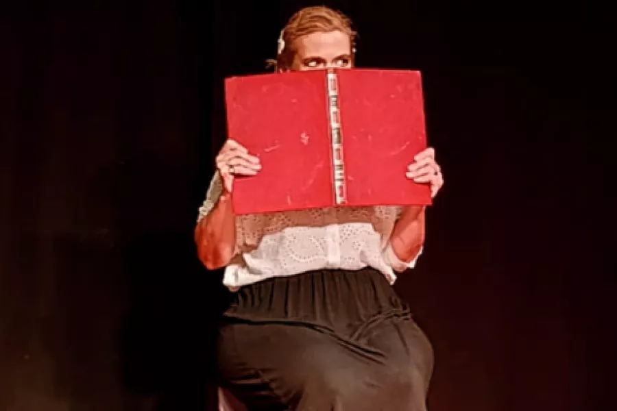 Es la imagen de una actriz caracterizada como Alfonsina Storni, escritora argentina. Está vestida con una camisa blanca y una pollera negra y está sosteniendo una carpeta roja que le tapa la boca