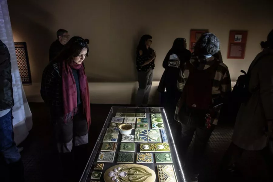 En primer plano una vitrina exhibiendo diversos modelos de azulejos, alrededor visitantes observan atentamente.