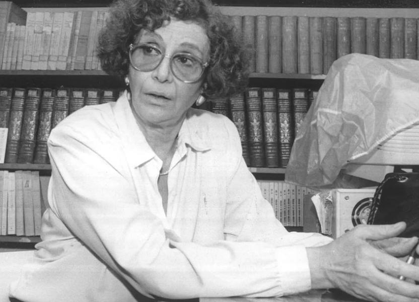 Hilda Habichayn sentada con las manos juntas teniendo una lapicera. Una biblioteca repleta de libros en el fondo. 